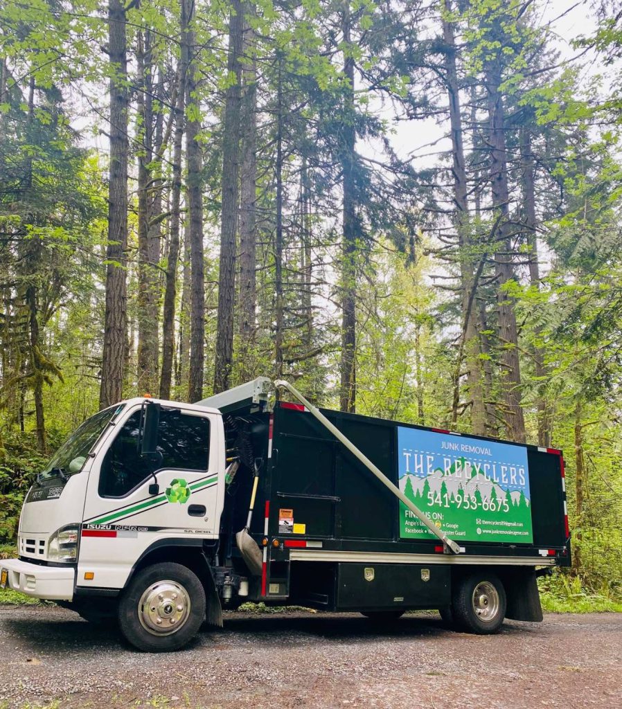 The Recyclers junk removal truck parked on a gravel road among tall Douglas firs